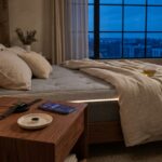 Nightstand with a smartwatch on a charger, a sleep ring on a tray, a smartphone showing a sleep app, a white noise machine, and a corner of a smart mattress in warm evening light.