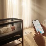 SNOO Smart Sleeper bassinet in a modern nursery with a swaddled sleeping newborn and a smartphone showing the SNOO app