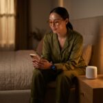 Person wearing amber blue light glasses in a dim bedroom holding a phone with a smart mattress and smartwatch visible on the bedside, warm lighting and a white noise machine on the nightstand